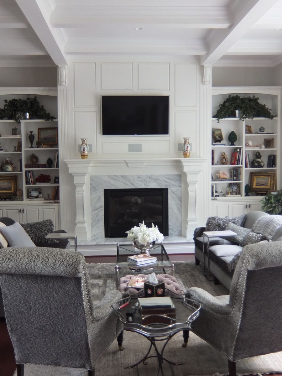 Clean, traditional renovated living room with white paneled walls and built-in shelves, gray furniture, and marble fireplace